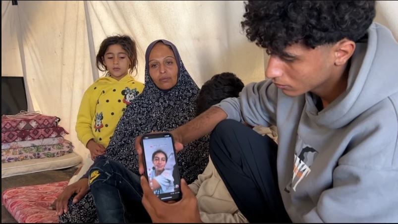 مردی در حال نشان دادن تصویر دخترش روی موبایل در کنار یک زن و کودکی در یک چادر در غزه
